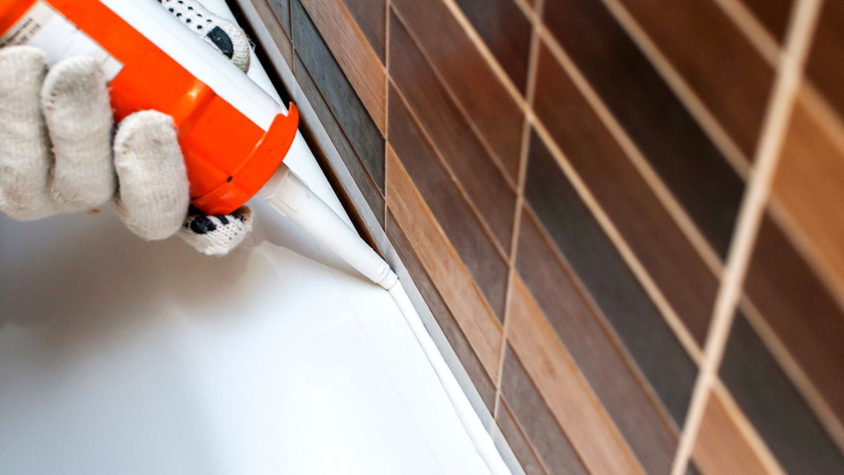 Sealing a bath with silicone and a caulk gun next to brown tiles