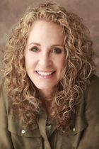 a headshot photo of ADHD coach Cheryl Susman: a woman with blond curly hair wearing a brown button up shirt against a lighter brown background smiling facing the camera