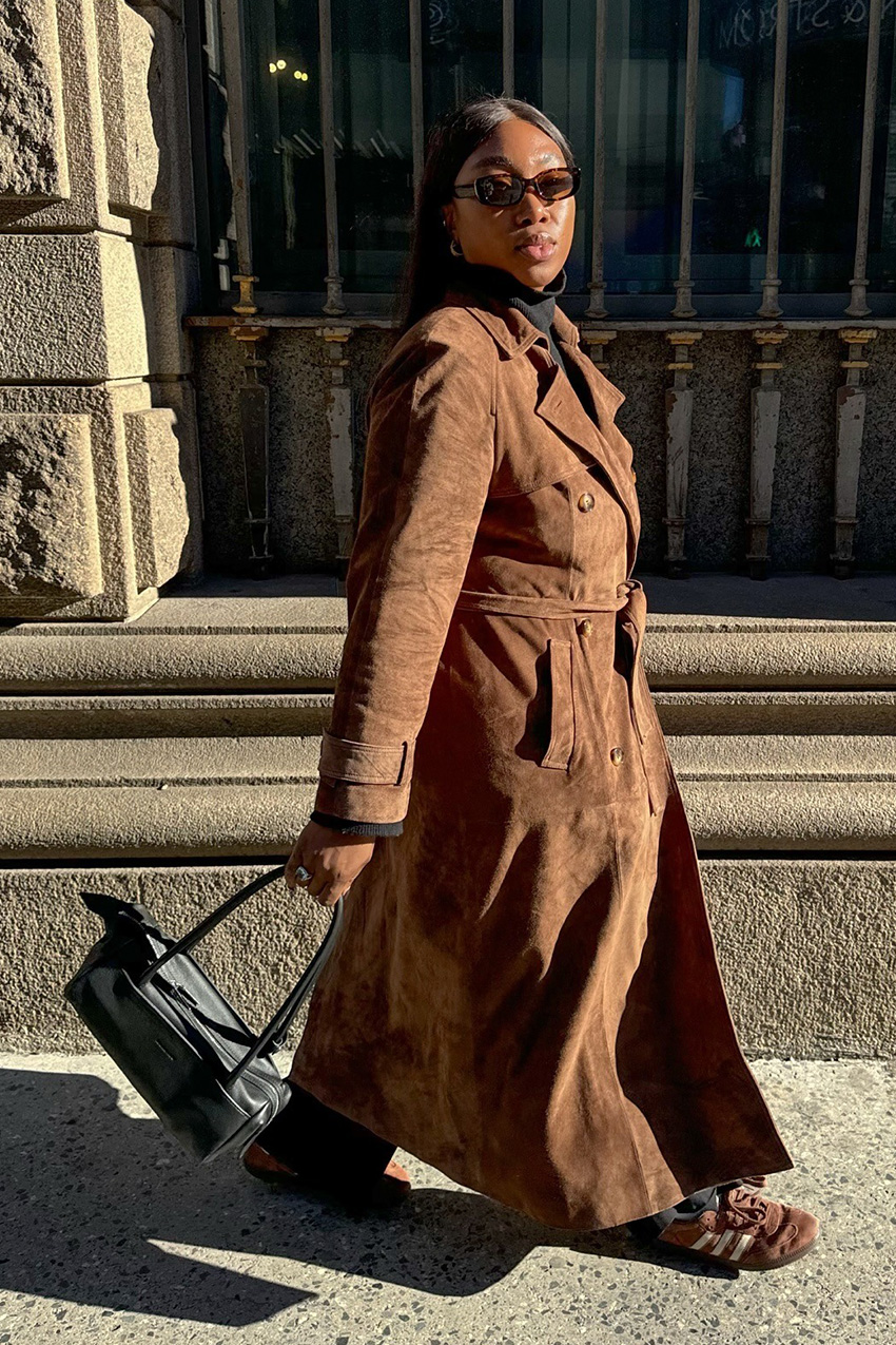 The best statement coats are shown in a street style photo of a woman walking down the sidewalk wearing brown sunglasses with a black turtleneck, a brown suede trench coat, black trousers, brown suede addias sneakers, and a black shoulder bag