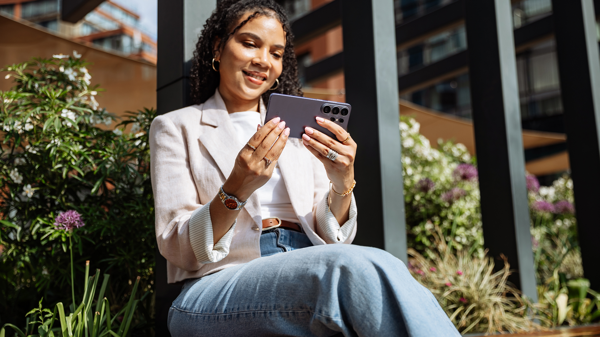 A woman sits outside, smiling at her Samsung Galaxy S26 Ultra phone, which she holds horizontally in both hands