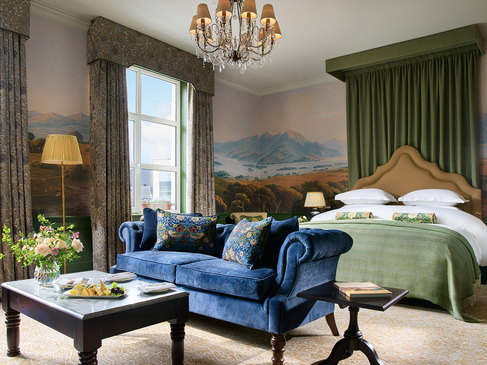 A luxurious hotel suite with a blue sofa and bed with green curtains behind it