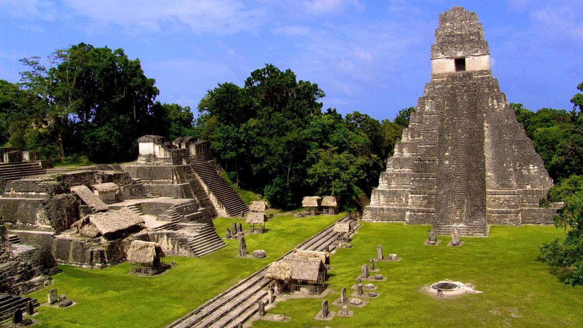 Maya ruins of Tikal in the jungle of Guatemala. On the right a tall step pyramid, with a series of stone steps leading to a doorway at the very top. To the left are two smaller step pyramids, half the size of the larger step pyramid. There are several rows of gravestones dotted around the green lawns. There are tall, lush trees in the background.