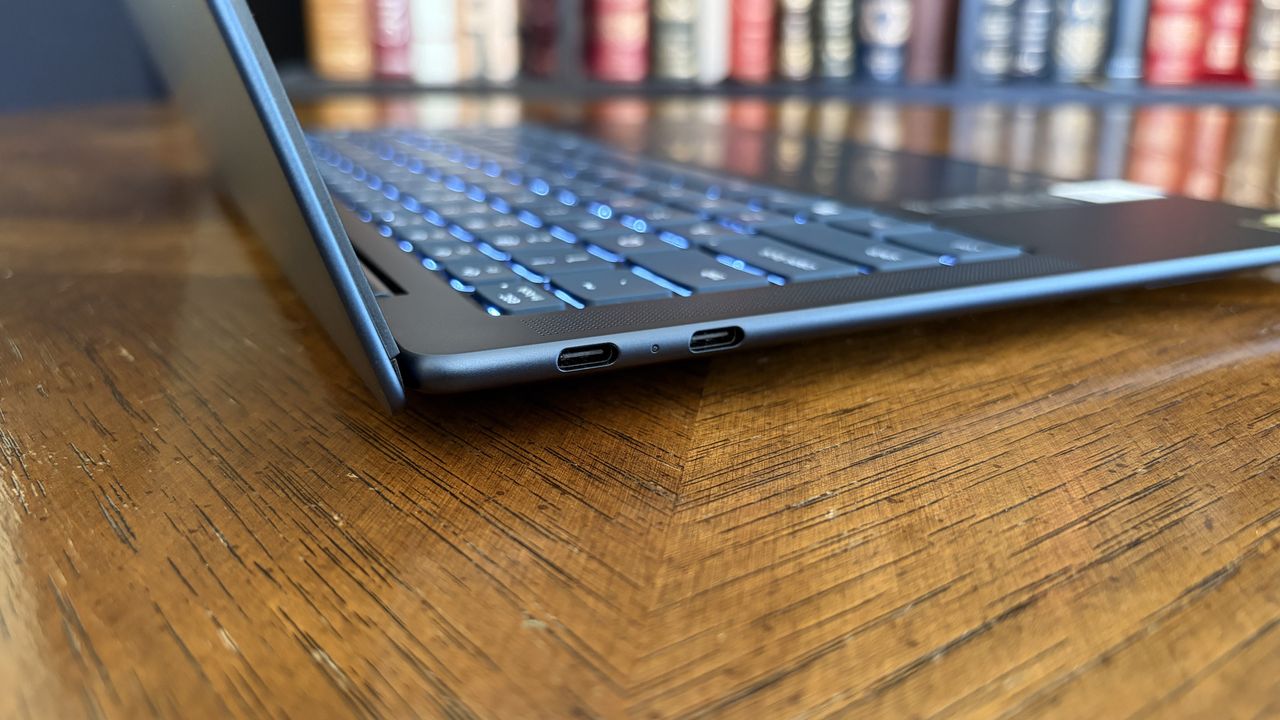 A sleek, open laptop on a wooden table focuses on its USB-C ports. Background shows colorful, blurred books on shelves, suggesting a studious setting.