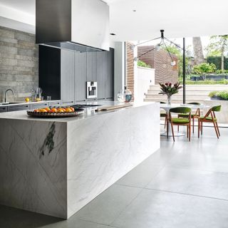 Kitchen with grey tiled concrete floor, large marbled kitchen island, floor to ceiling glass doors to the patio and garden