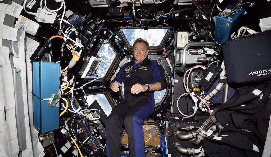 a man in a blue flight suit poses inside the international space station, with earth in the background