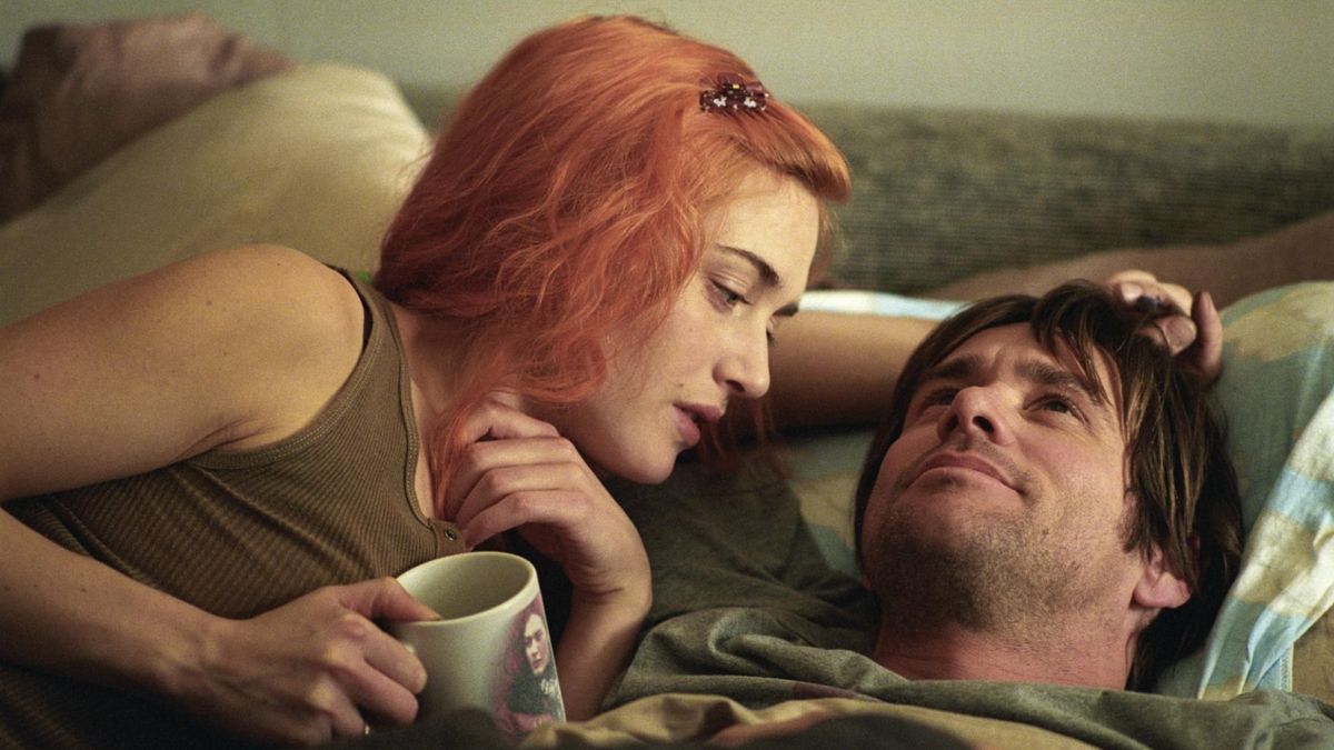 A young couple lying a bed together. The girl has red hair and is holding a coffee cup while looking at the dark haired man, who is smiling and looking up at the ceiling.