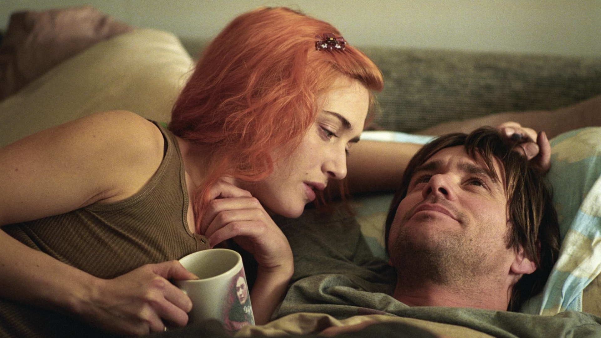 A young couple lying a bed together. The girl has red hair and is holding a coffee cup while looking at the dark haired man, who is smiling and looking up at the ceiling.