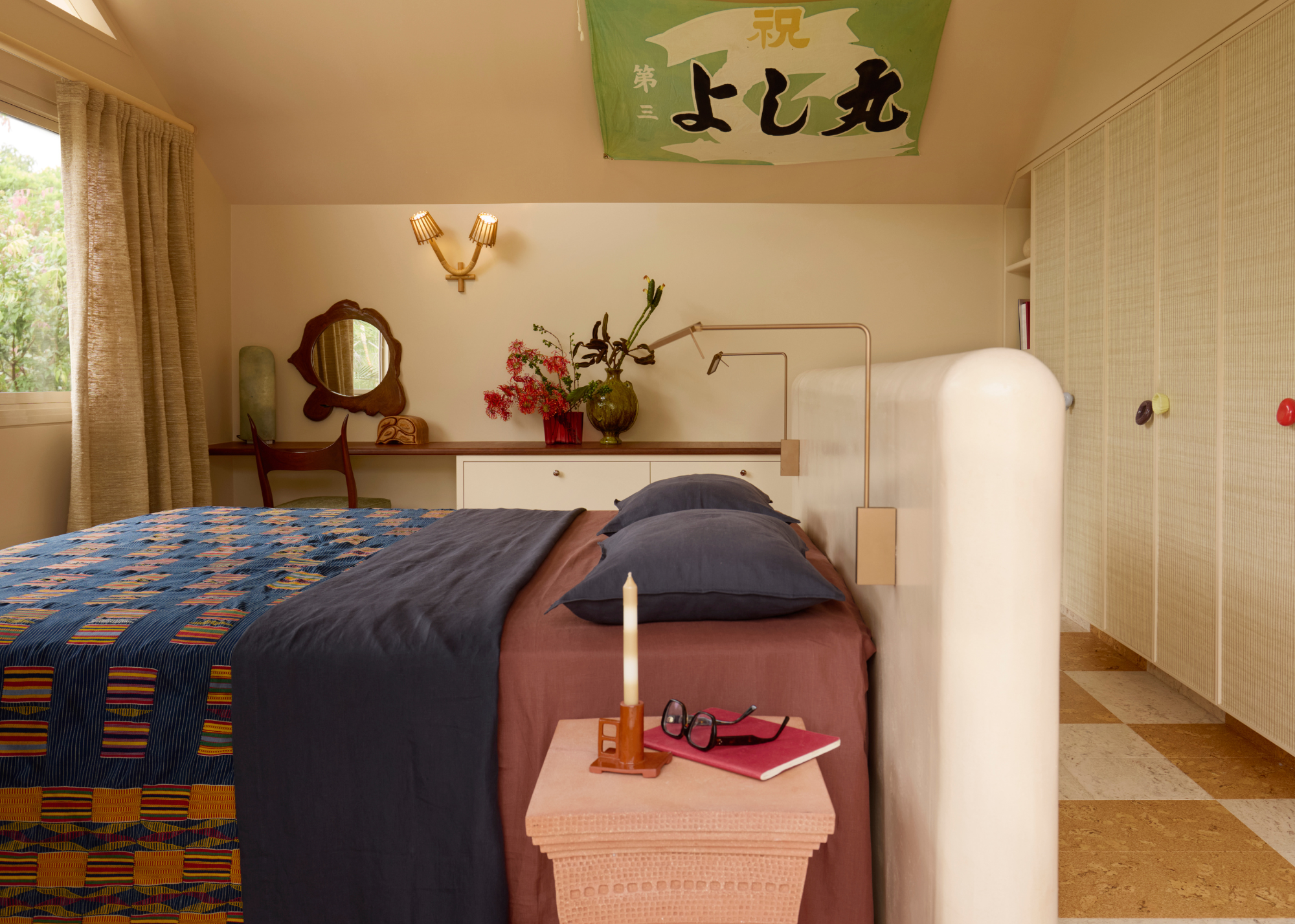 A modern bedroom with the bed in the center of the room with a built-in resin headboard creating a division between the fitted wardrobes behind; there is a tapestry hanging from the angled ceiling and a built-in ledge against the back wall used as a dressing area and decorative display