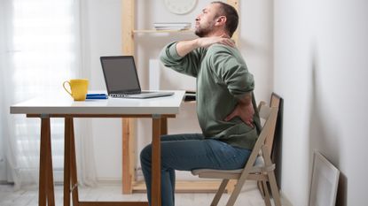 man sitting sideways to the camera on a chair, at a desk with a laptop on. he's holding his side and shoulder as though in discomfort. 