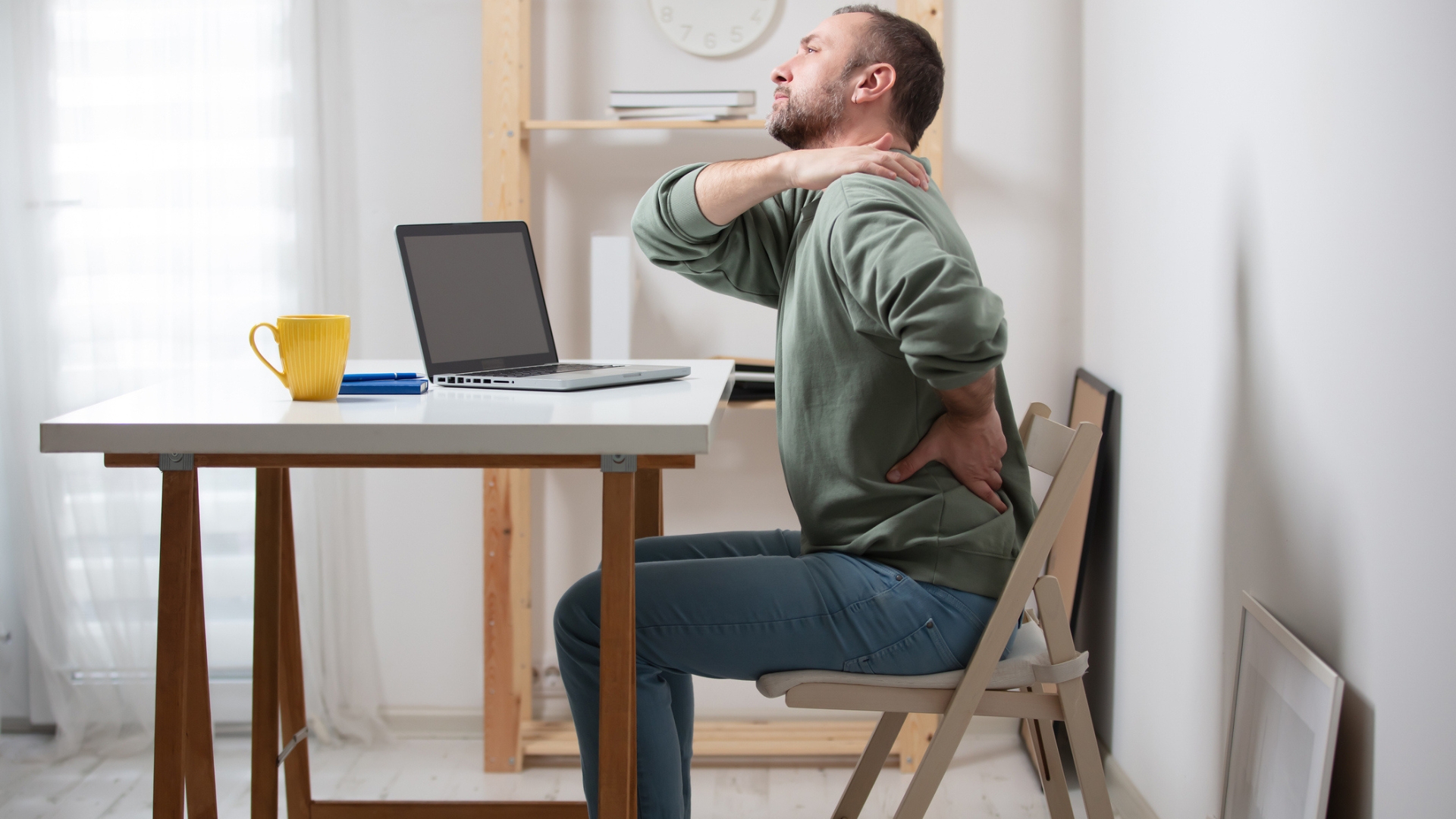 man sitting sideways to the camera on a chair, at a desk with a laptop on. he&#039;s holding his side and shoulder as though in discomfort. 
