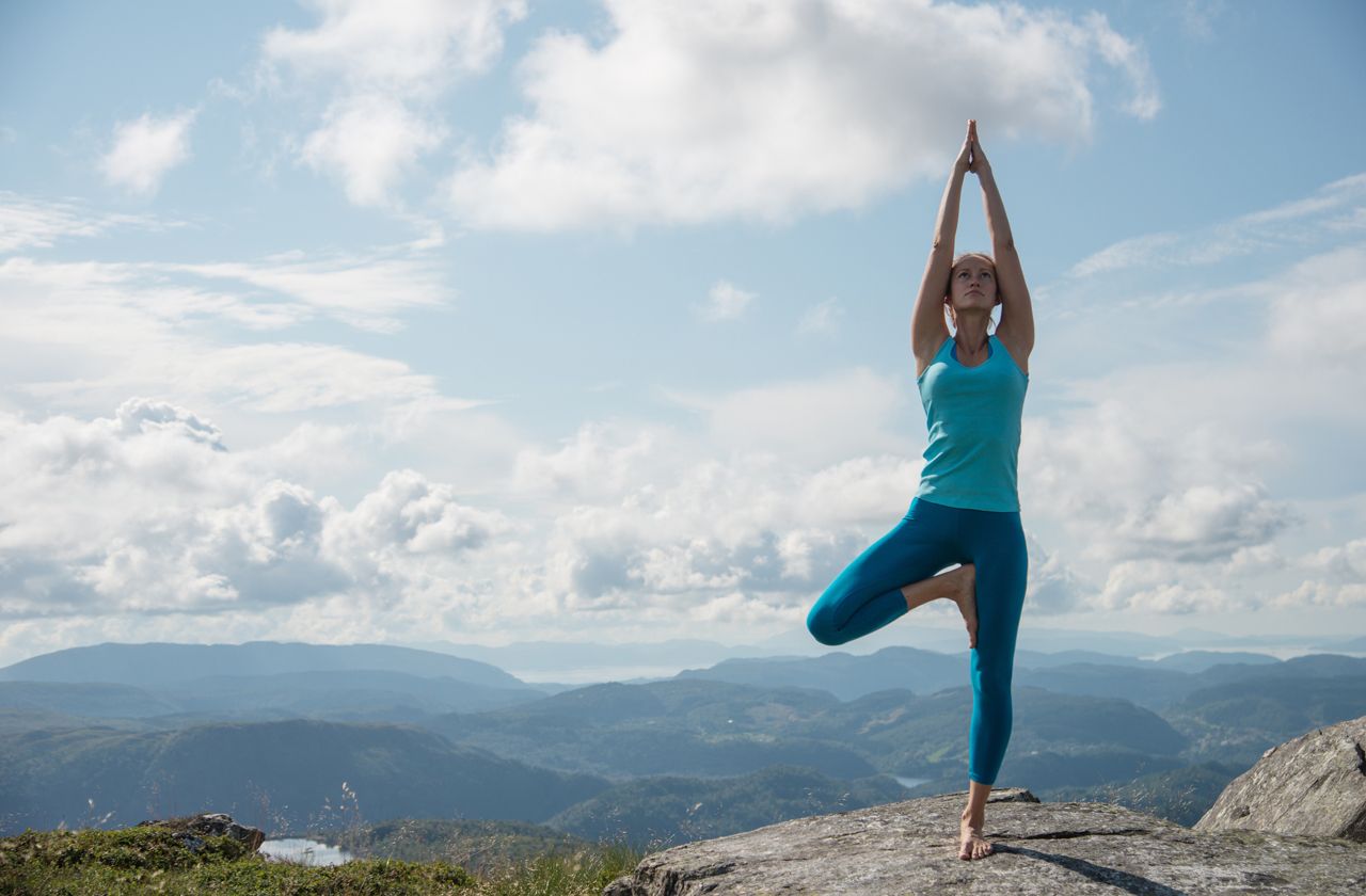 Young woman practicing yoga high up on top of mountain Ulriken, in Bergen, Norway, overlooking mountain range - tree pose or Virksasana.