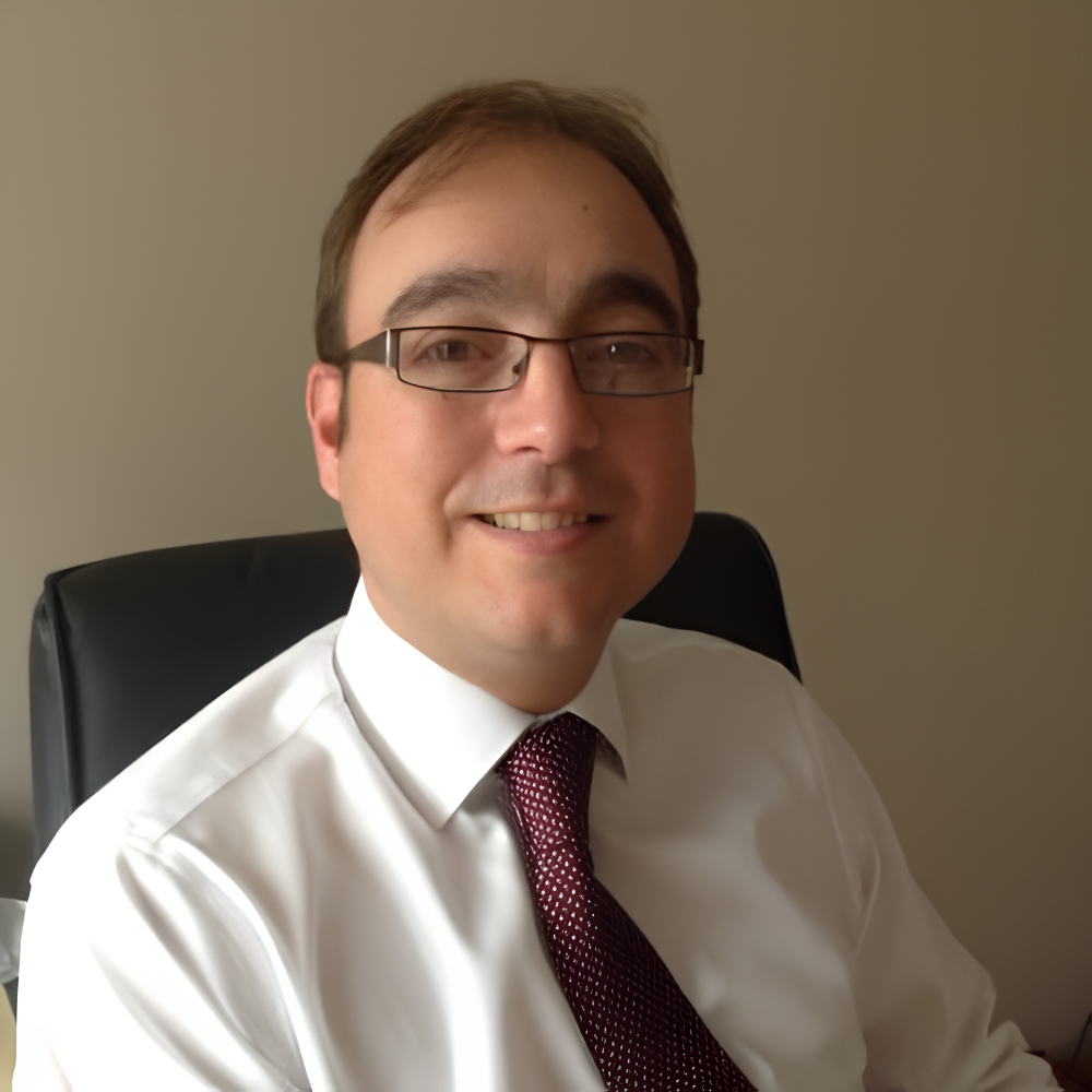 Man with glasses and short brown hair, smiling in a white shirt and maroon tie, seated in an office chair.