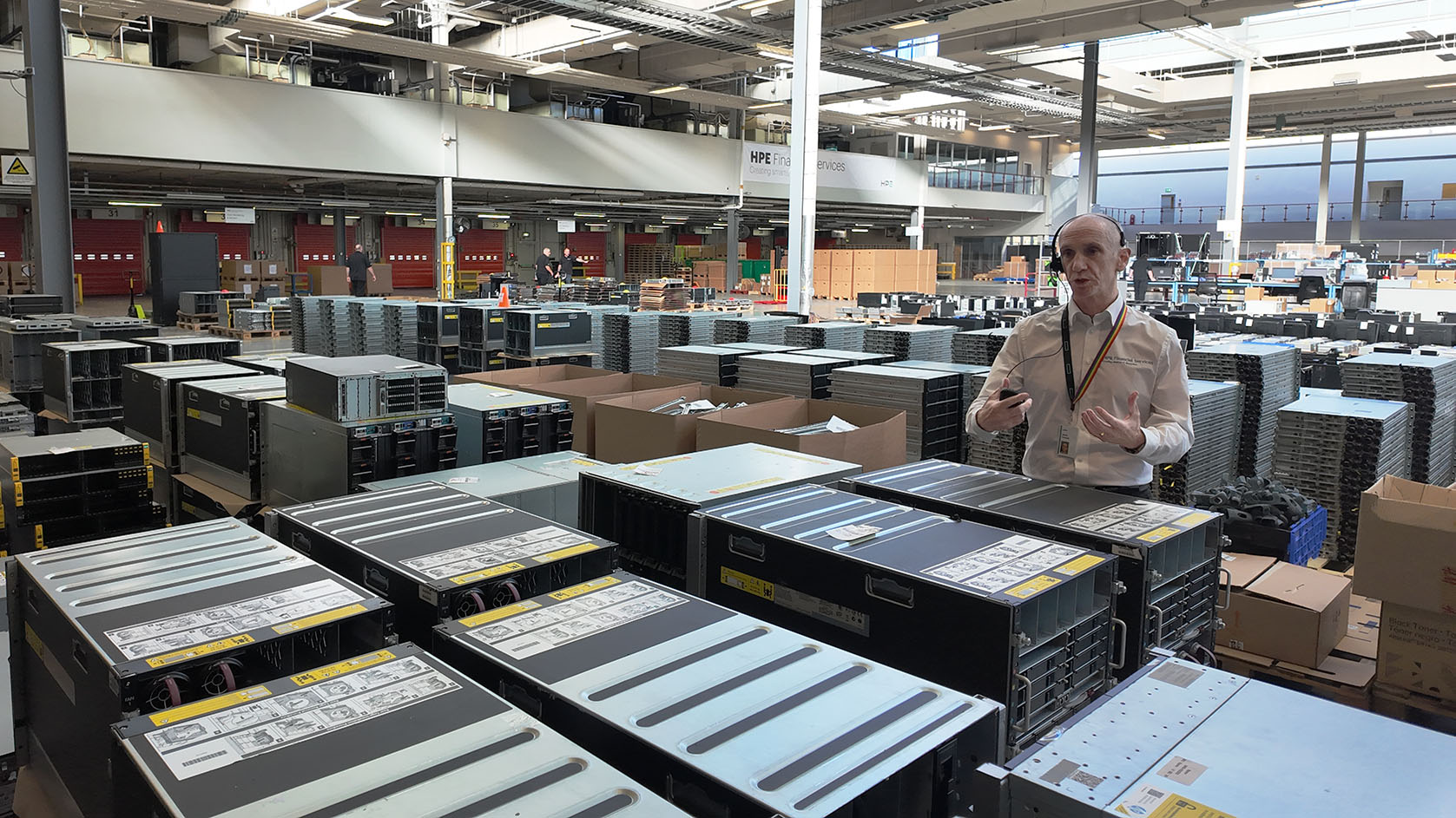 A man in a white shirt and a headset stands in between rows and rows of carefully piled up used hardware including PC towers and servers.