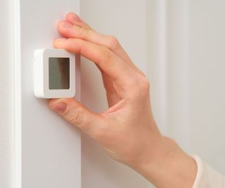 A Caucasian hand adjusting a small white square hygrometer on a white wall