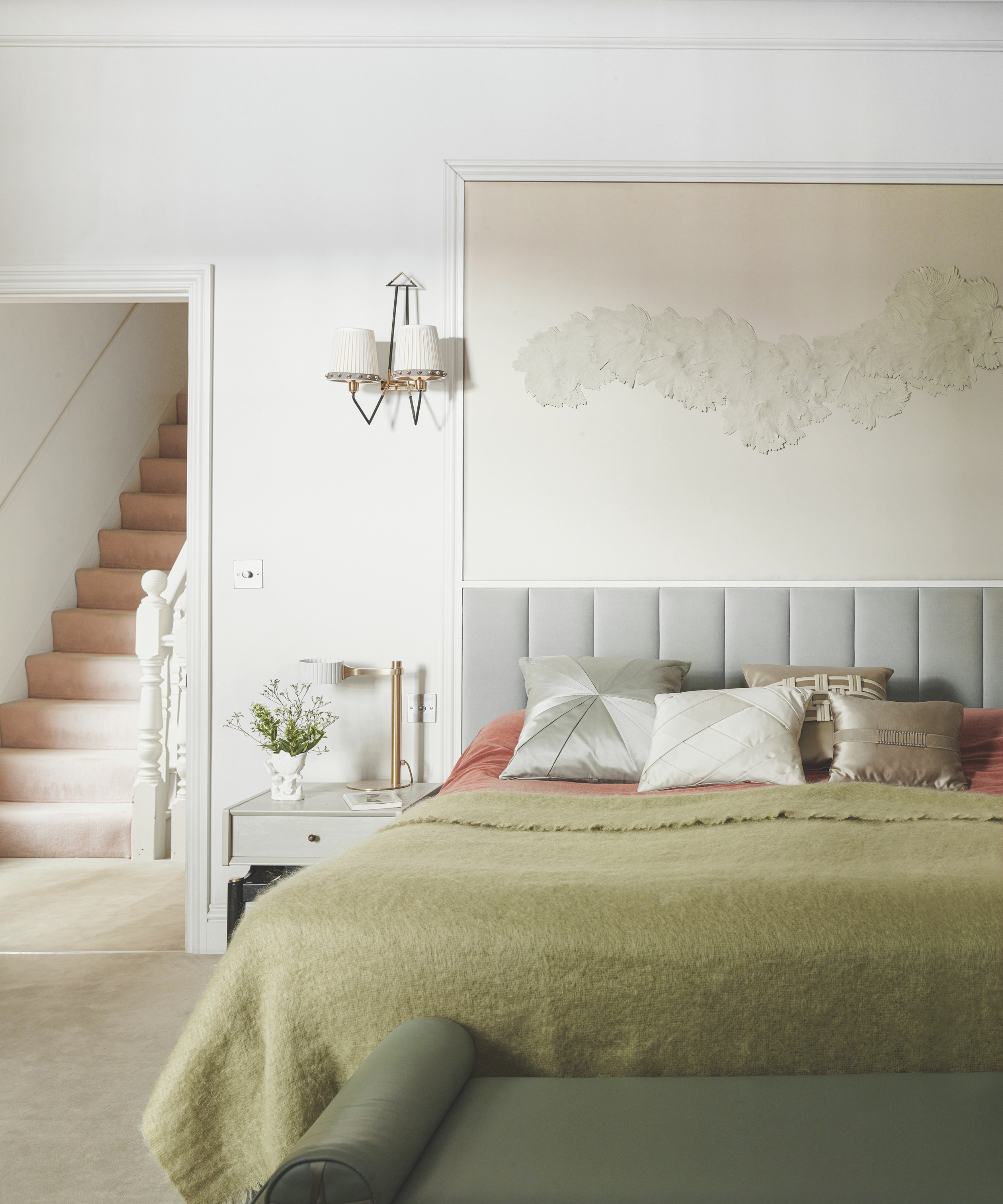 a calming bedroom with a green blanket on the bed, lots of pillows, white artwork on the wall, a small side table next to the bed, and a sconce on the wall
