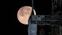A white rocket nose with the circular NASA logo stands next to the metal gantry underneath a glowing large full moon in a black night sky