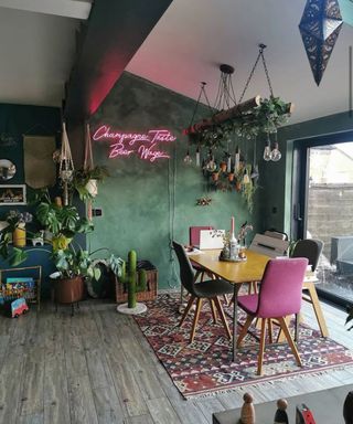 Quirky, jungalow style dining space with pink neon light sign, boho patterned rug, pink and gray mix and match chairs, and hanging industrial lights with greenery overhead.