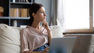 A young woman sitting on a sofa at home with a laptop on her lap looking thoughtfully into the distance