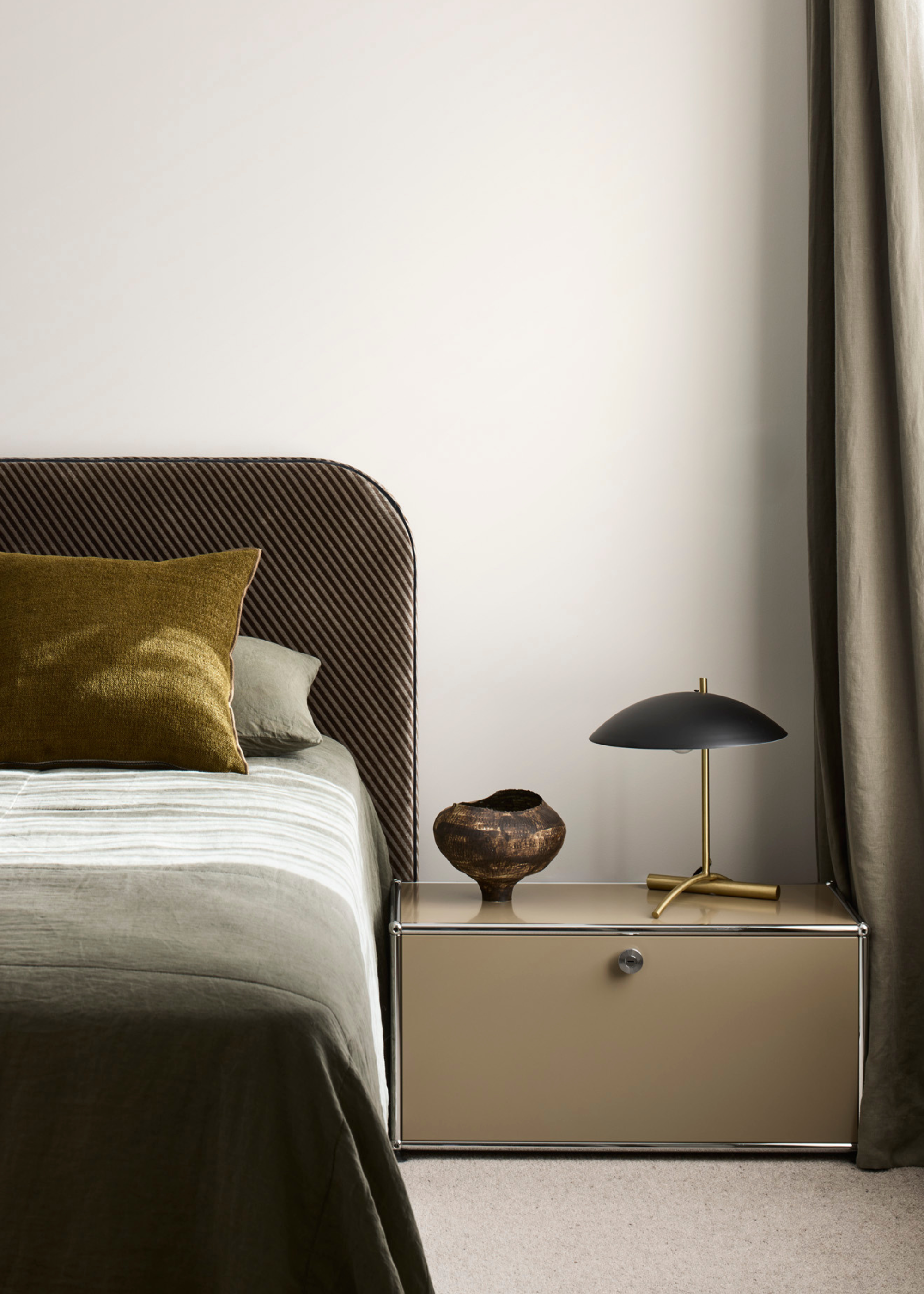 A white bedroom with a curved headboard, a green pillow, a nightstand with a vase and a table lamp