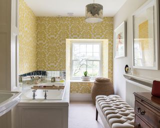 Elegant bathroom with traditional basin and bath, ottoman and antique chest of drawers, yellow and white wallpaper and large laundry basket.