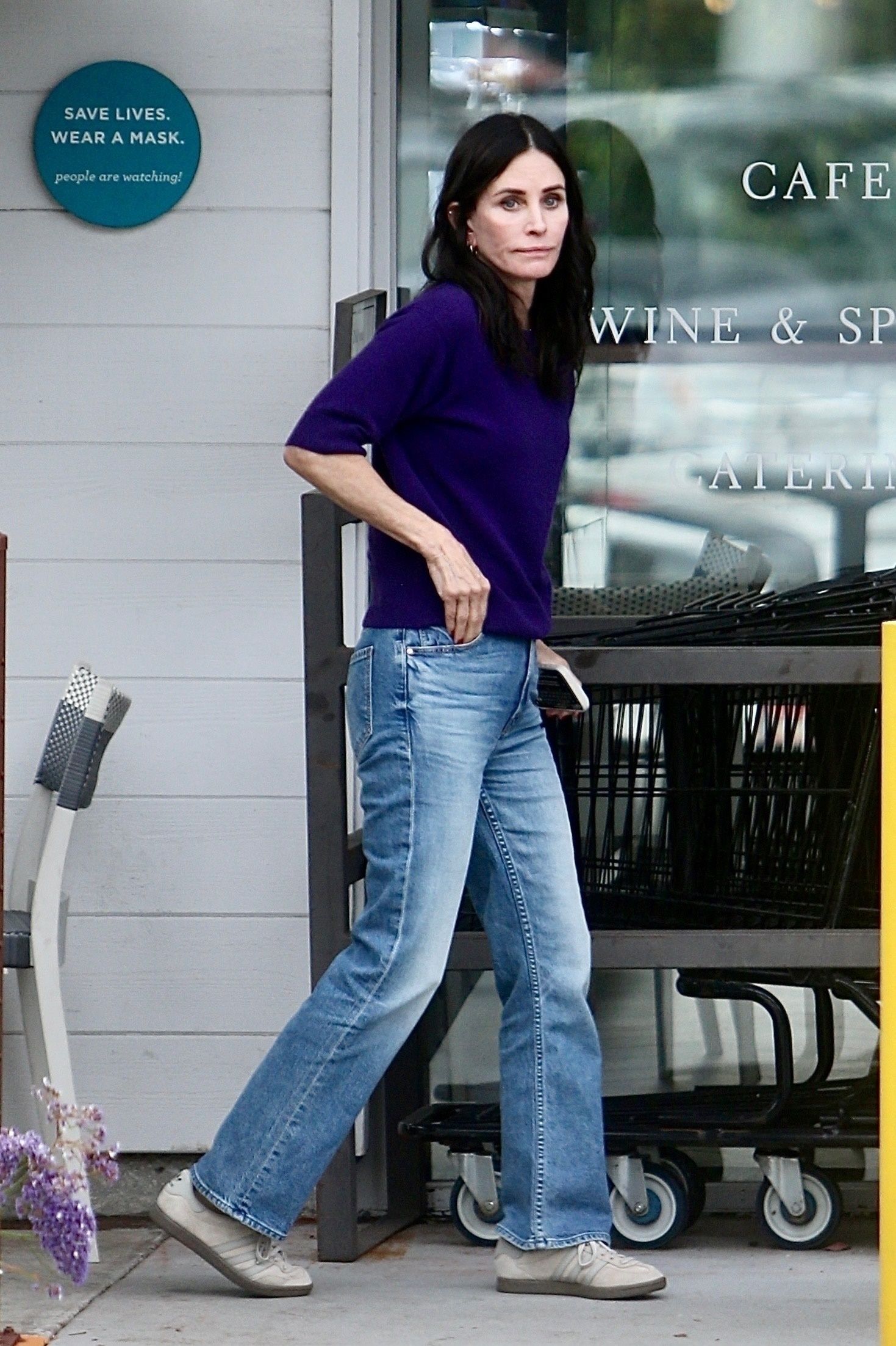 Courtney Cox walks outside wearing light grey trainers with slim-fit jeans and a dark purple knit.