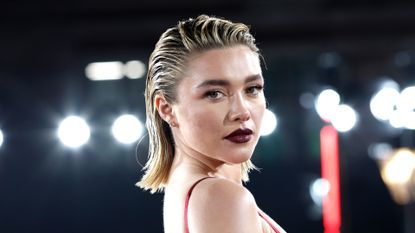 Florence Pugh has slicked back blonde hair with dark roots and wears a dark red lipstick and a red strappy dress