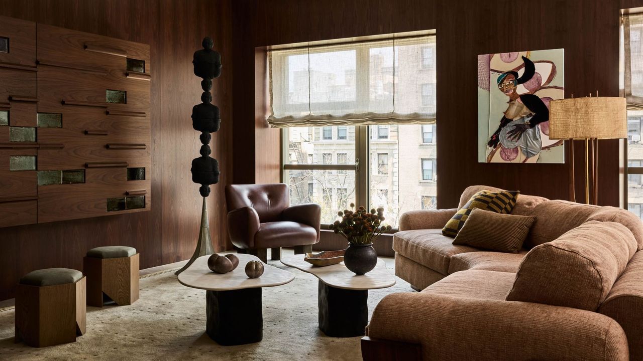 An art collector&#039;s loft living room with brown walls. structural sculptures, a brown couch, upholstered wooden pouffs, a pair of curved wood coffee tables, and a brown leather armchair