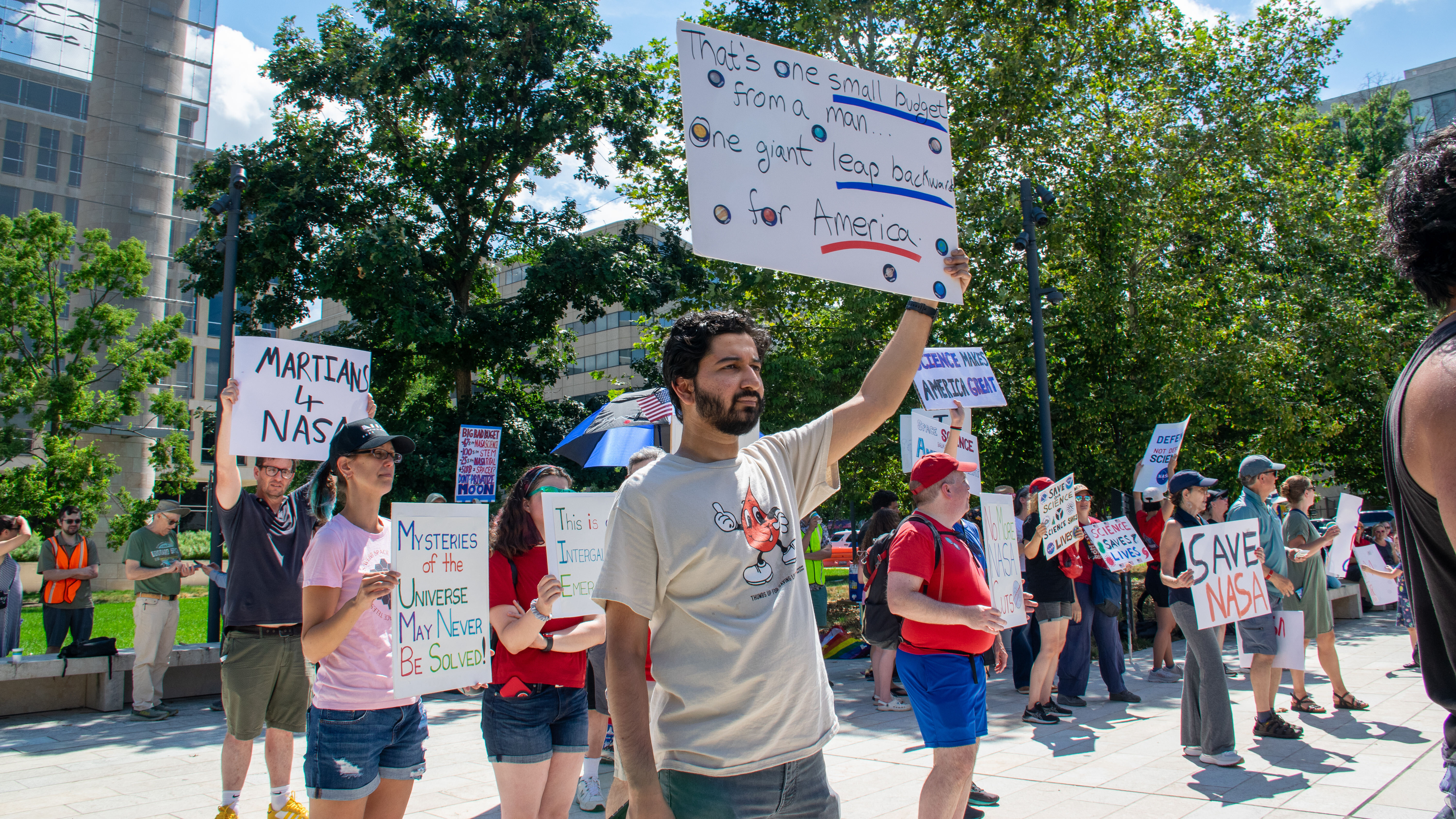 People hold signs outside on a sunny day in protest of NASA budget cuts, July 20, 2025.