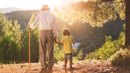 A grandpa walks with a cane in a sunlit field with his grandson.