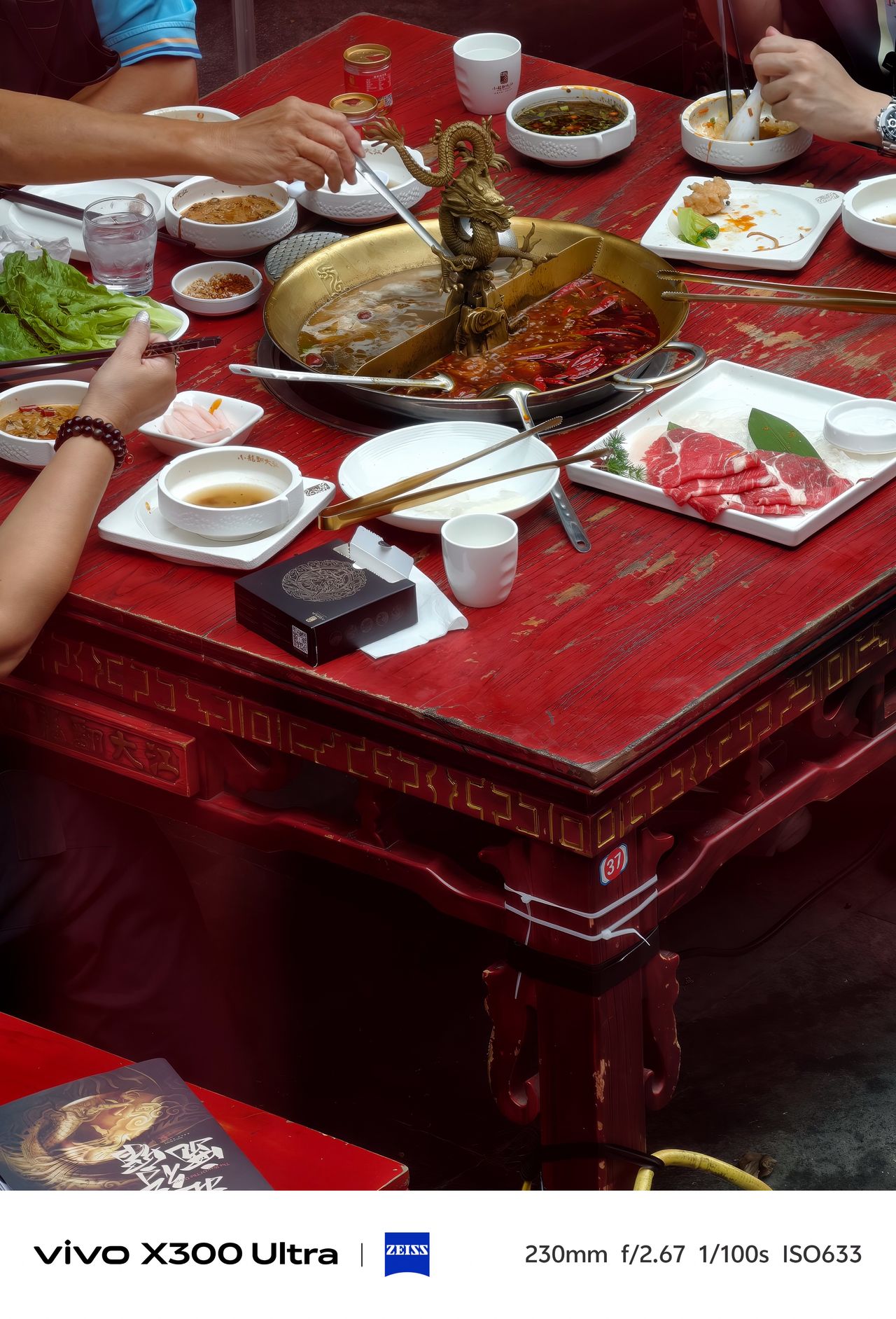 Diners using chopsticks around a split brass hot pot container on a red wooden table.