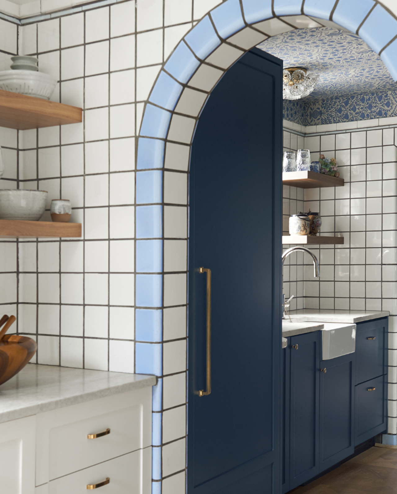 Image of a kitchen with white cabinetry and white tile walls. The door reveal is a light blue tile and the doorway leads into a room with dark blue cabinetry and a farmhouse sink. There are floating wood shelves in the room.