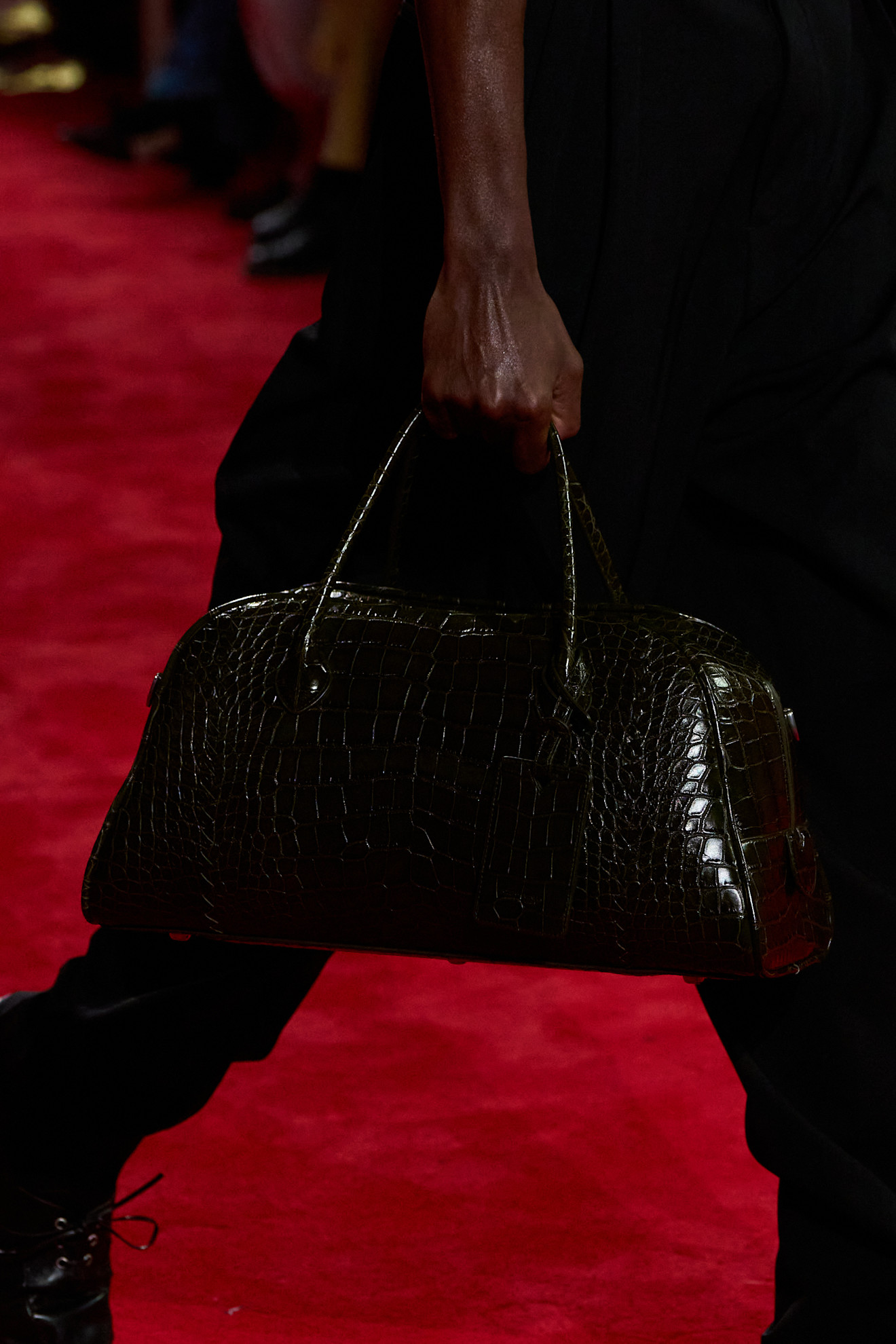 A close-up photo of a model walking in the F/W 26 Bottega Veneta show carrying a dark-green croc top-handle bowler bag.