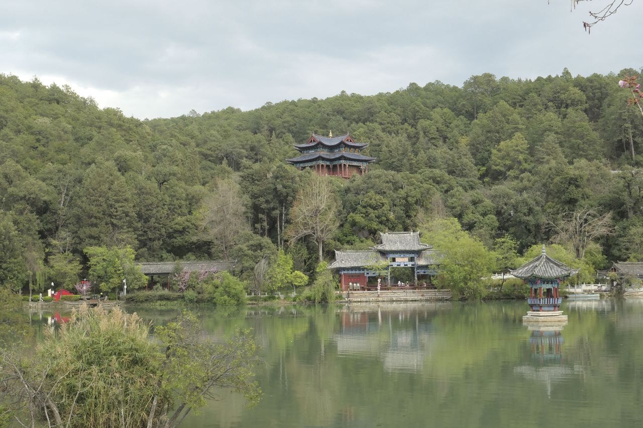 A Chinese temple nestled among trees, with a still lake in the foreground