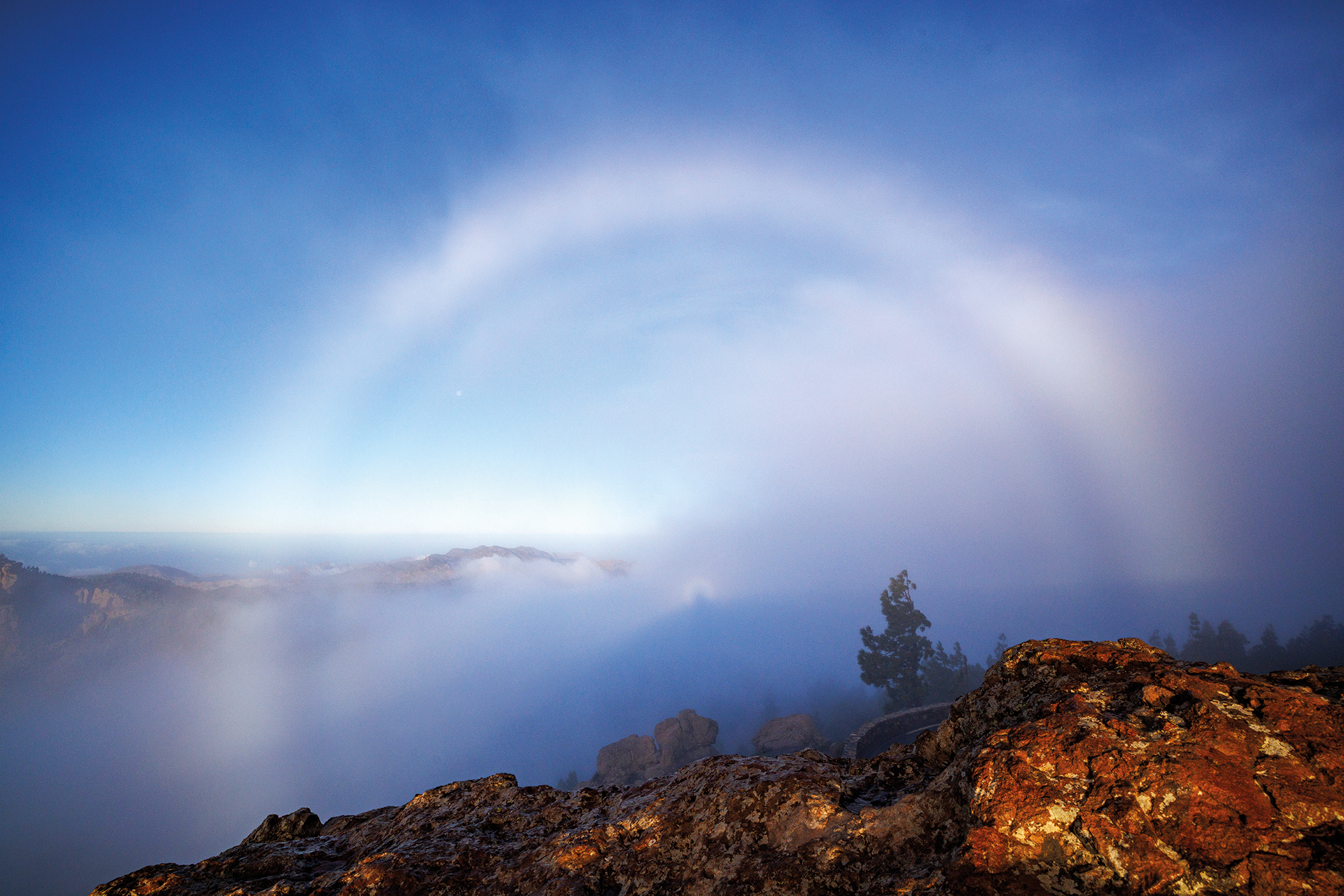 A misty white rainbow arches over a rocky mountain peak amidst fog and soft, blue sky, evoking a serene and mystical atmosphere