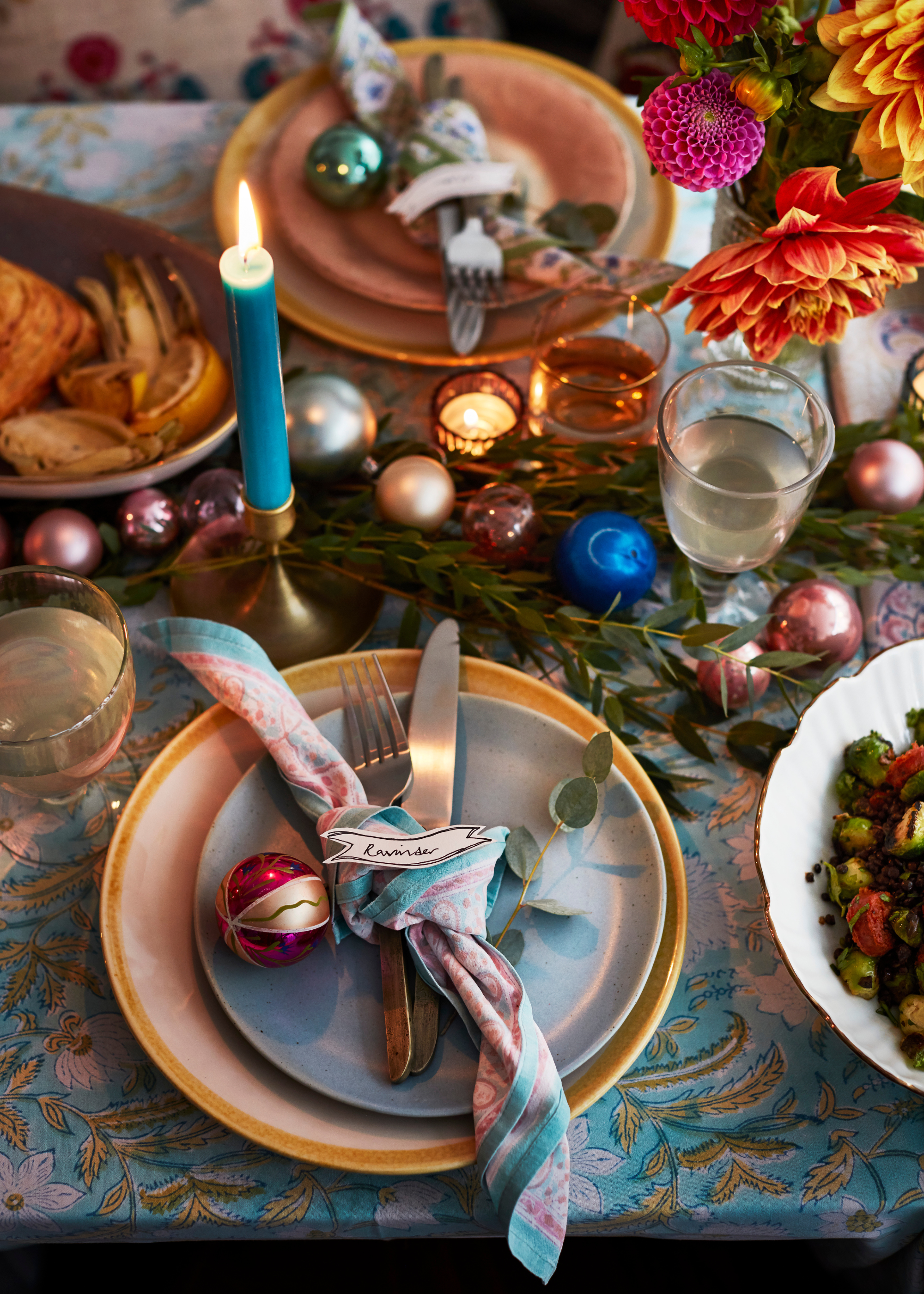 A Christmas table set for a meal with a tied napkin and bauble on each plate, and extra baubles, foliage, taper candles and tealights scattered across the table with colorful flowers in a vase