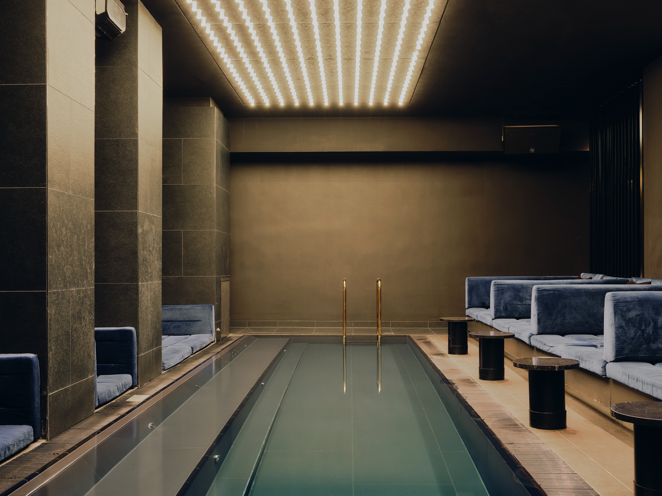 A spa pool with rows of LED lighting above the water and velvet or suede blue sofas along either side of the pool's edge with round dark wood tables on the side without the pillars