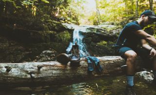 En man sitter på en stock framför ett litet vattenfall i skogen, med vandringsskängorna och strumporna placerade på stocken.