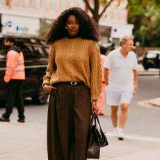 Woman wears caramel sweater, brown trousers, and Tabi flats while carrying a handbag and shopping bag. 