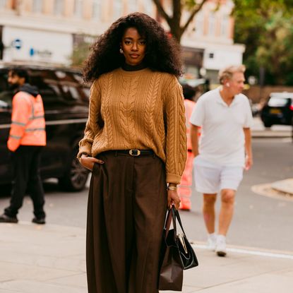 Woman wears caramel sweater, brown trousers, and Tabi flats while carrying a handbag and shopping bag. 
