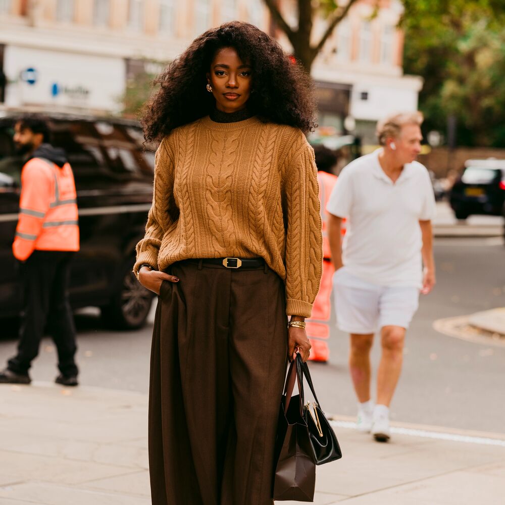 Woman wears caramel sweater, brown trousers, and Tabi flats while carrying a handbag and shopping bag. 