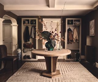 square hallway with round table in centre, cream rug, dark grey walls and storage units to rear of room either side of a white door