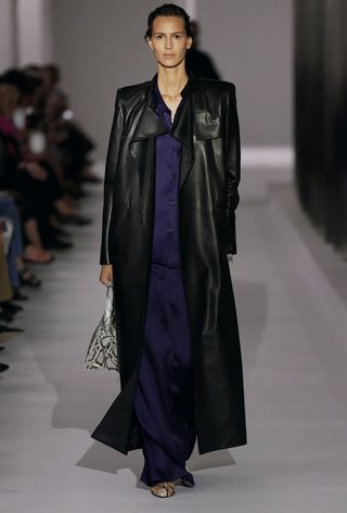 A snakeskin purse is shown in a photo of a model walking down the of Khaite's spring 2025 runway show carrying a python handbag with a a long black leather trench coat, navy blue satin shirt, navy satin trousers, and snakeskin heels