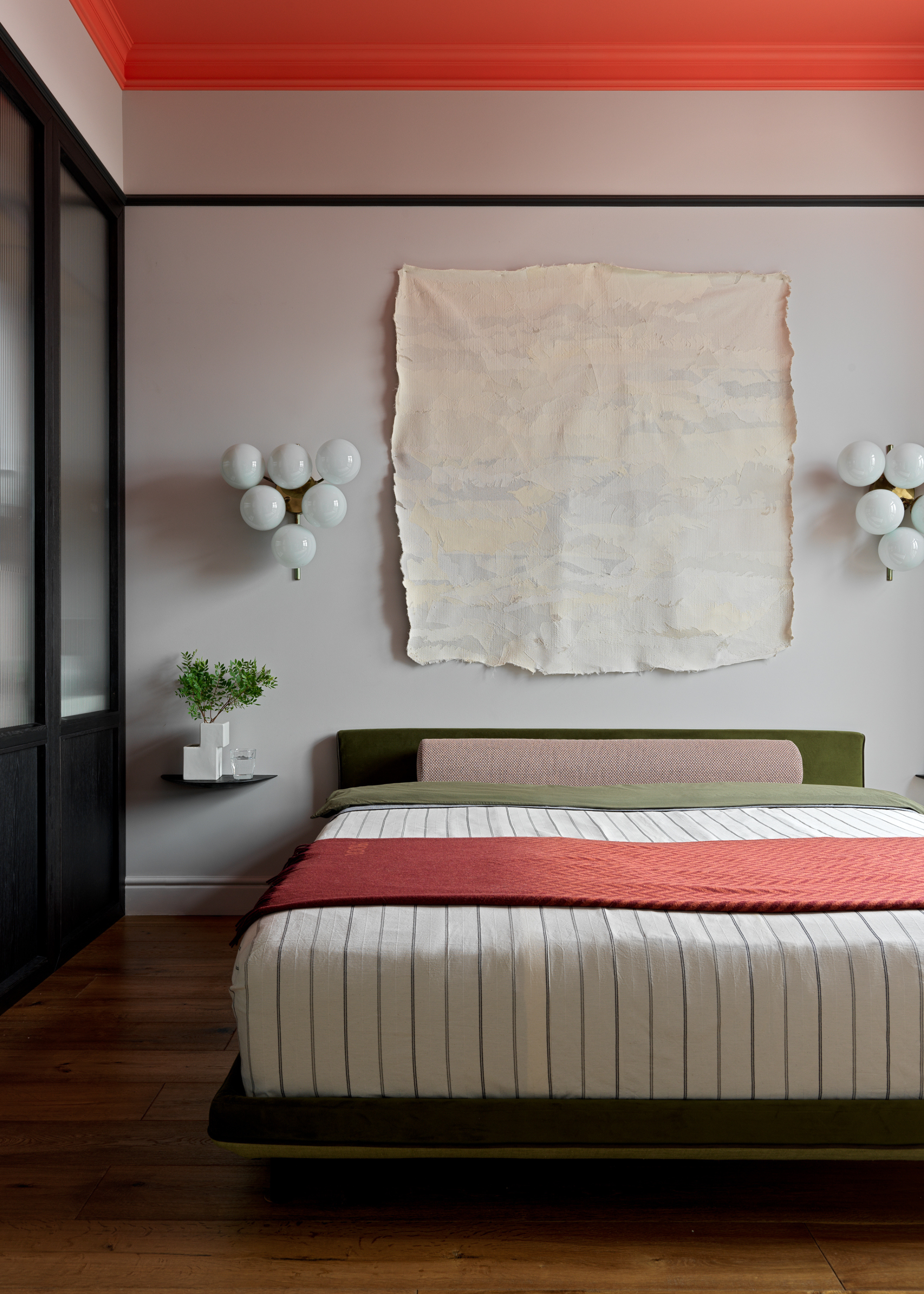A white bedroom with clustered wall lamps, a tattered painting, floating side tables with vases, a low bed with a striped bedscape, and a brown bolster pillow