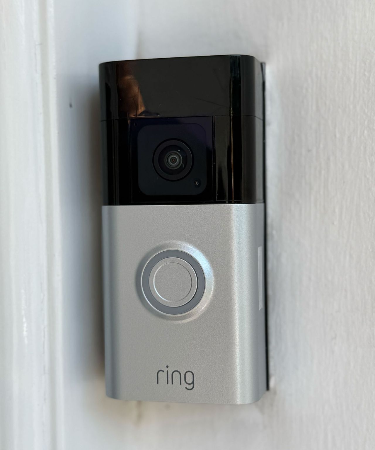 A black and silver Ring Battery Doorbell Plus mounted on a white painted wall. It features a camera lens, circular button, and Ring logo.