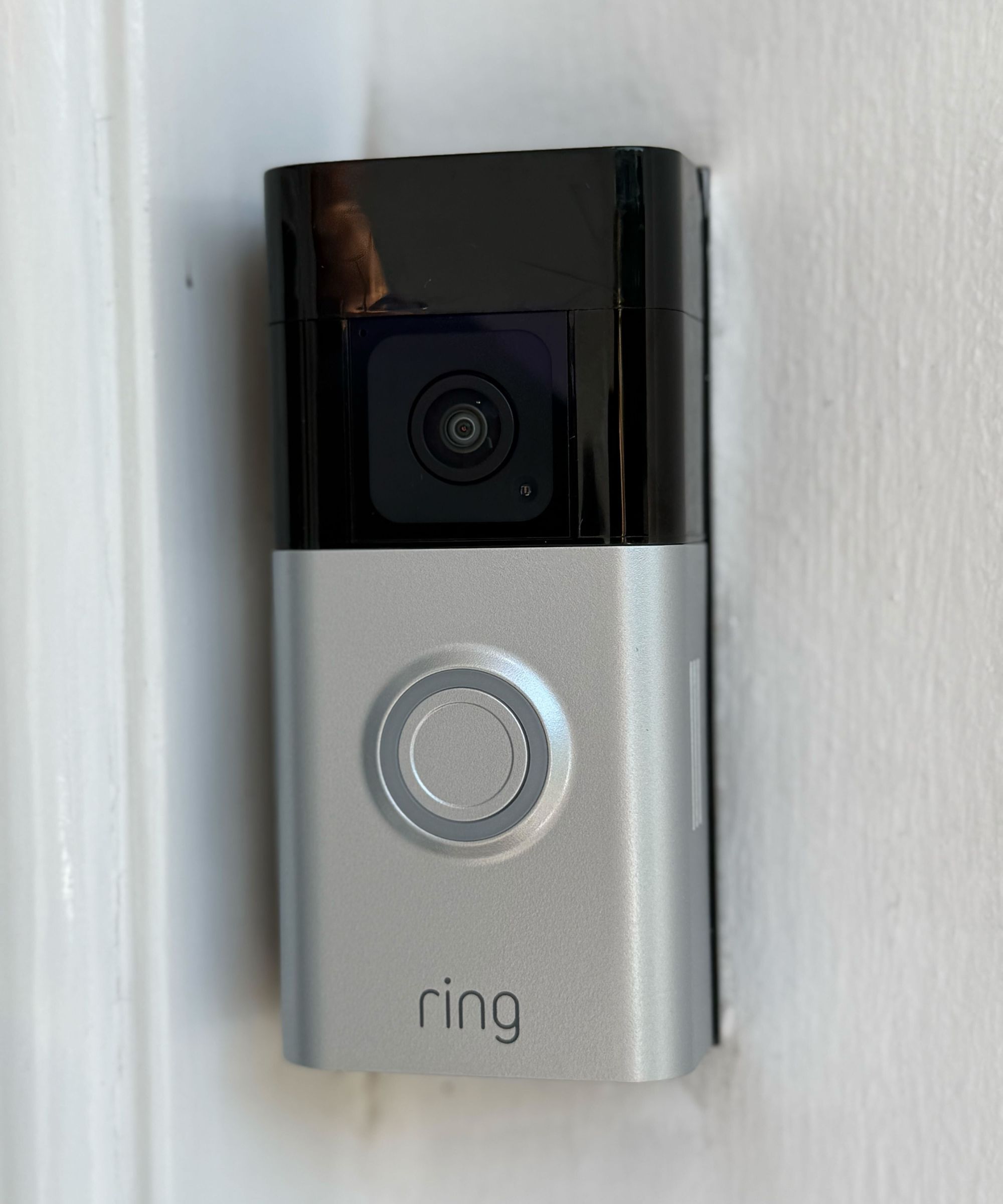 A black and silver Ring Battery Doorbell Plus mounted on a white painted wall. It features a camera lens, circular button, and Ring logo.