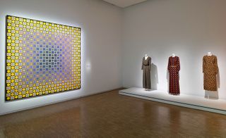View of Centre Pompidou's tribute to Yves Saint Laurent featuring three mannequins displaying patterned fashion pieces and colourful patterned wall art in a room with white walls and wood flooring