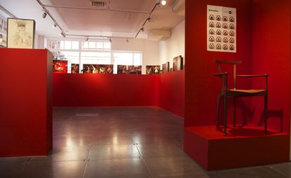 Display room, slate grey floor, white wall and ceiling with spotlights, windows letting in light far wall, red wall surround display with female erotic art along the top, wooden stool on a red viewing platform with artwork above on a red wall front of shot