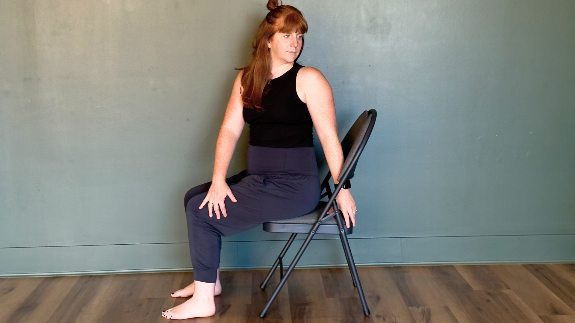 woman performing a seated twist on a chair on a wooden floor against a green wall