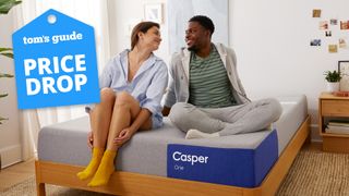 Image shows a happy couple chatting and sitting on the Casper The One memory foam mattress placed on a wooden bedframe in a white bedroom, with a blue Tom's Guide price drop deals badge overlaid on the image in the top left hand corner (left)