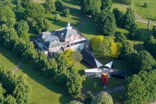 In a lushly green park, a futuristically designed pavilion featuring a star-shaped structure extends from the entrance of a brick-shaded palatial building.